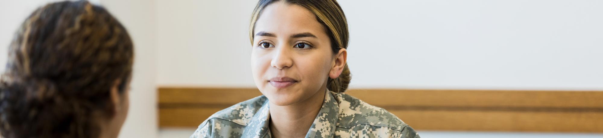 A female military veteran wearing fatigues sits across a table from a woman in civilian clothes