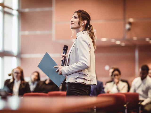 business people listen to a woman speaking at a conference