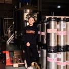 Tapping Potential scholarship winner Dung Ngô stands alongside canisters of beer and other brewing equipment. She wears a black sweatshirt  that reads "Furbrew" in orange lettering.